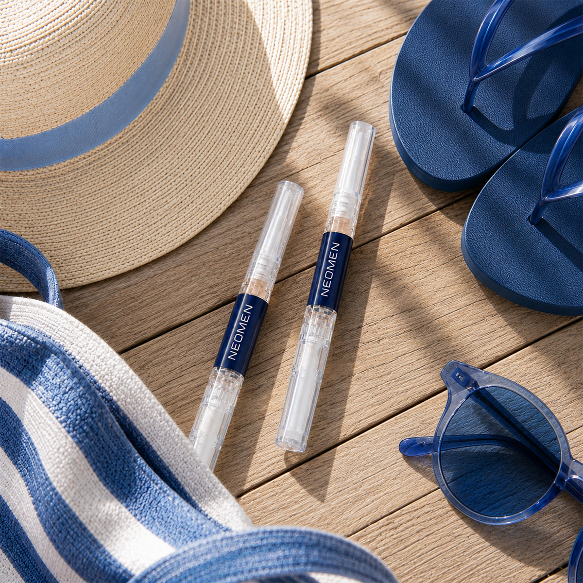 Two Neomen nail renewal pens on a wooden deck with a straw hat, sandals and sunglasses